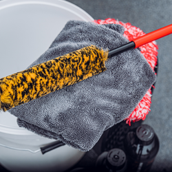 A yellow and black cleaning brush rests on a folded gray microfiber towel near a white bucket, with red and black cleaning supplies visible in the background.