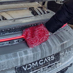A person wearing a black sleeve uses a red microfiber mitt to wash the snowy rear of a Porsche car with a VDM Cars license plate.