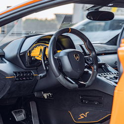 Close-up of a Lamborghini sports car interior, featuring a black and yellow dashboard, sporty steering wheel, and orange accents. The exterior of the orange car and part of another vehicle are visible outside.
