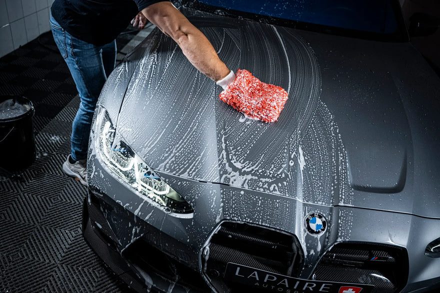 A person uses Best Summer DEAL to wash the hood of a gray BMW, creating soapy suds. The car’s headlights are on, and a black bucket sits nearby.