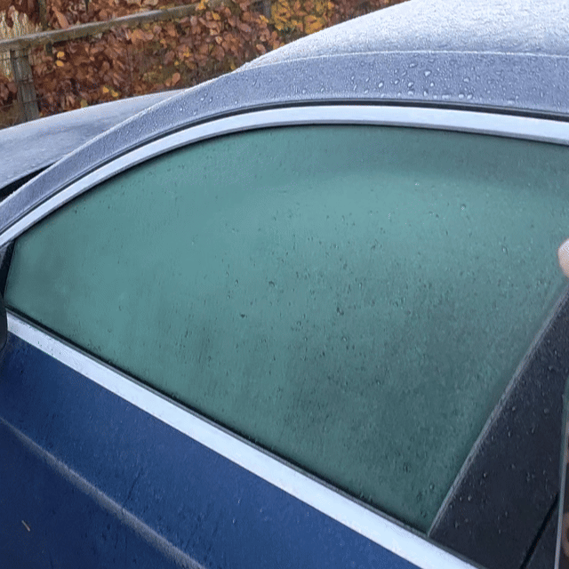 A car window frosted with condensation signals a chilly morning. Brown autumn leaves and a fence blur in the background—perfect timing to grab De-Icer for clear, safe driving.