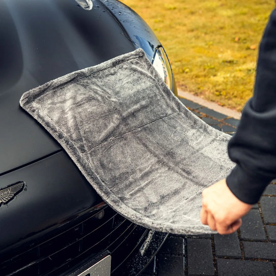Een persoon legt een grote grijze hoofddrooghanddoek op de motorkap van een zwarte auto die geparkeerd staat naast een grasveld.