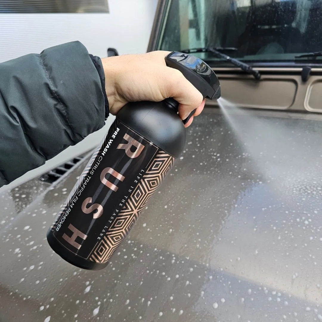 A hand sprays Pre Wash, a powerful traffic film remover, onto the wet hood of a car. The person in a dark jacket cleans the vehicle as part of an effective pre-wash routine; part of the windshield is visible.