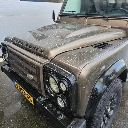 A brown Land Rover Defender with water spots on the hood and windshield is parked on a wet surface, ready for a Pre Wash using traffic film remover. The front grille, headlights, and part of the Dutch license plate are visible.
