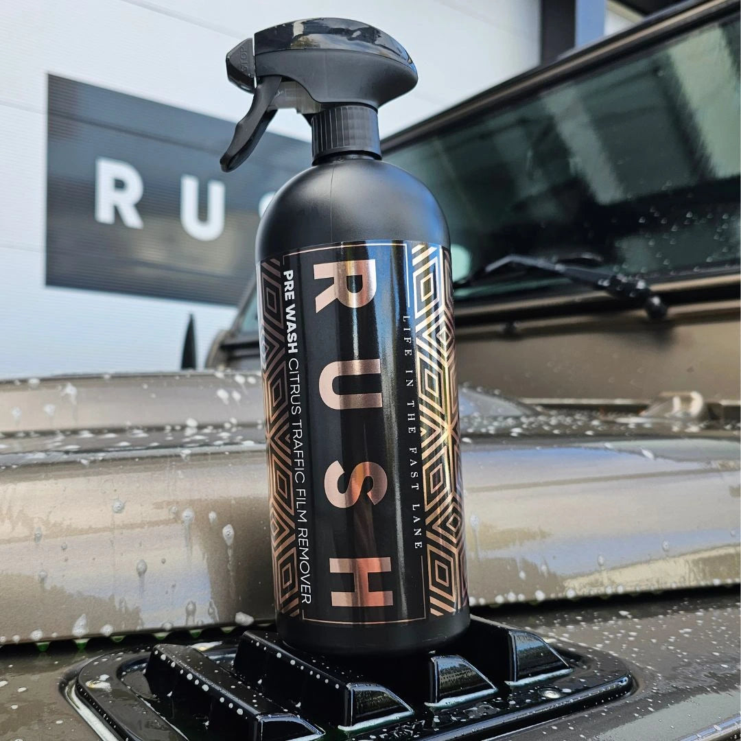 A black spray bottle labeled Pre Wash, an effective pre-wash auto alkaline cleaner, sits on the hood of a wet car with blurred RUSH signage in the background.