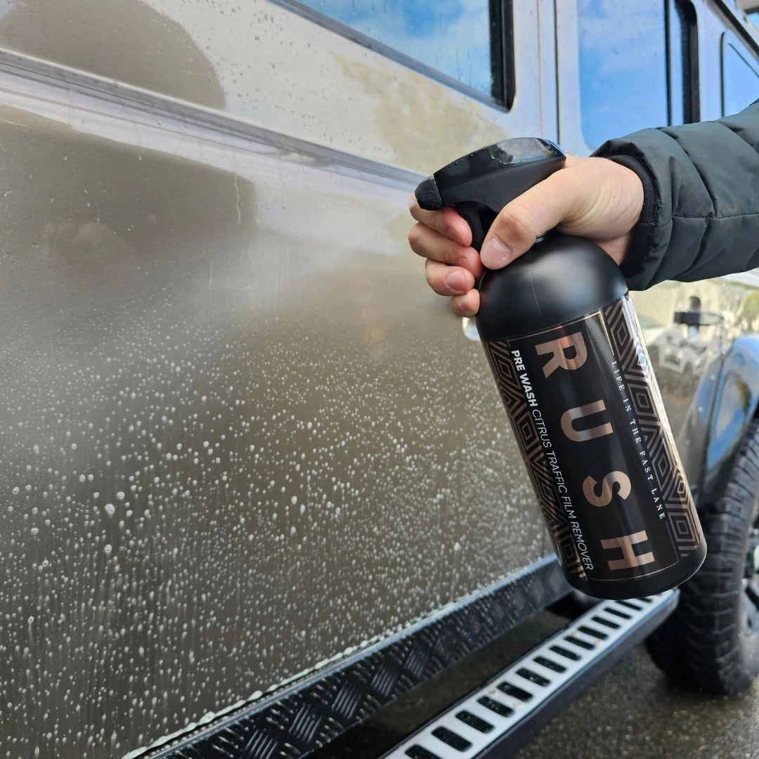 A gloved hand holds a bottle labeled Pre Wash, spraying foamy white bubbles onto the side of a beige car.