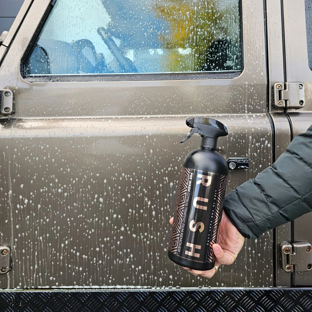 A person in a green jacket holds a black spray bottle labeled PUSH, applying Pre Wash to a soapy, partially cleaned car door.