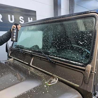 A hand sprays Pre Wash from a black RUSH bottle onto the soapy windshield of a brown car, with a building wall and RUSH sign visible in the background.
