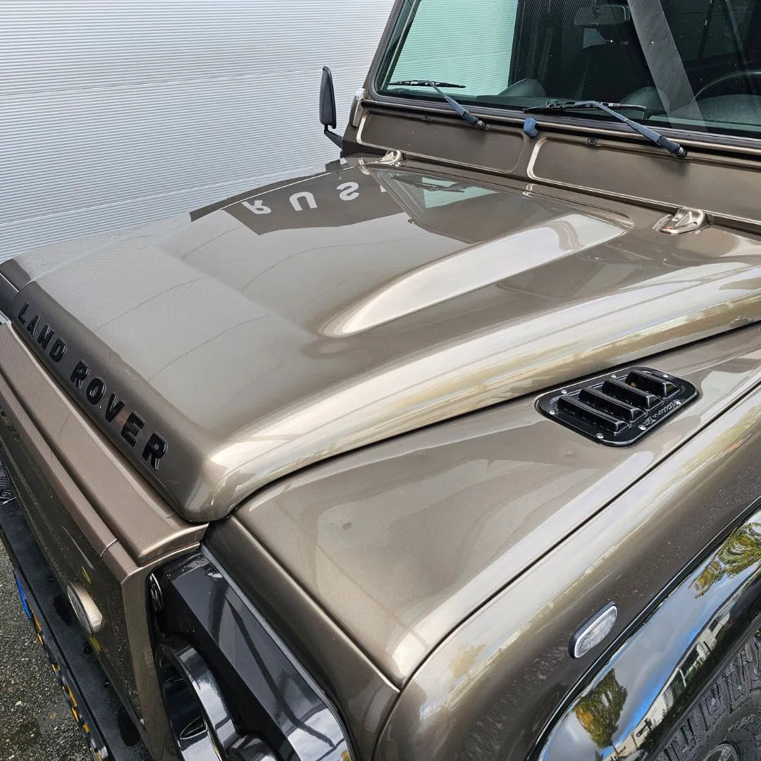Close-up of the front hood and windshield of a metallic bronze Land Rover, with the brand name on the hood and reflections on the shiny surface treated with 1+1 GRATIS Spray Wax for lak bescherming using nanotechnologie.
