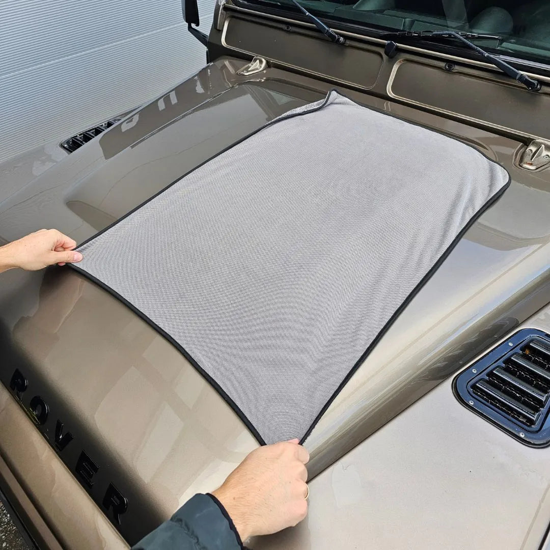 Twee handen spreiden een grote grijze microvezel droogdoek 600 GSM uit over de motorkap van een beige voertuig waarop ROVER letters zichtbaar zijn.