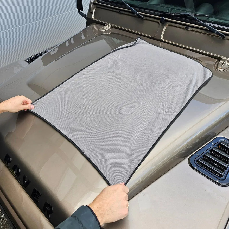 Twee handen spreiden een grote grijze microvezel droogdoek 600 GSM uit over de motorkap van een beige voertuig waarop ROVER letters zichtbaar zijn.