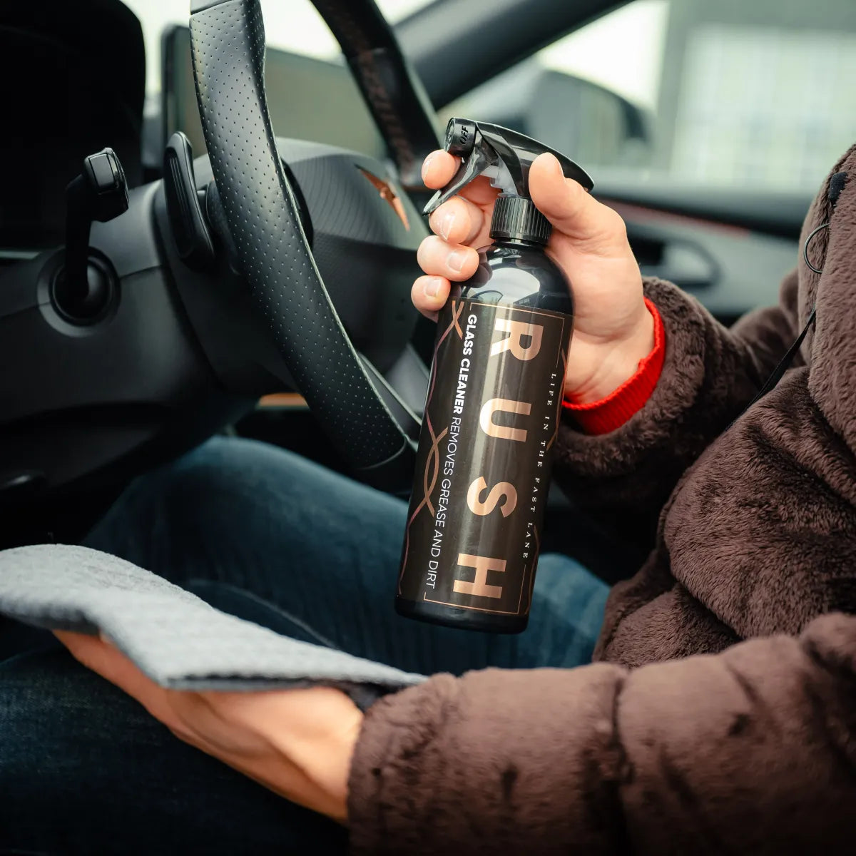 Een persoon in een auto houdt een RUSH Window Clear Winter Kit en een grijze doek vast en maakt het interieur schoon.