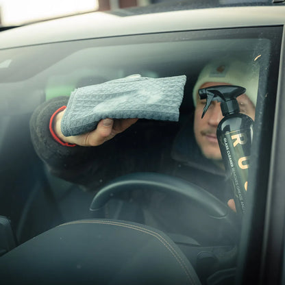 Een persoon in een auto reinigt de voorruit met een grijze doek, RUX spuitfles en de RUSH Window Clear Winter Kit.
