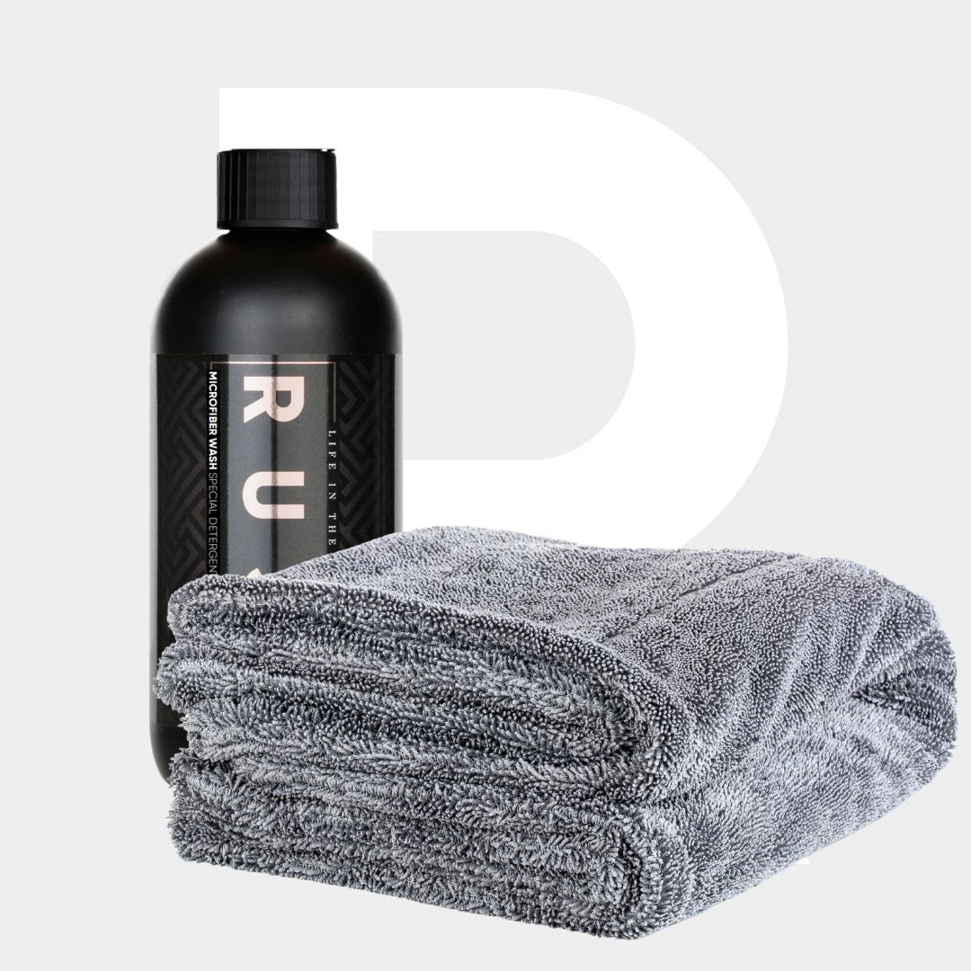 Een zwarte fles Microfiber Wash met een zwarte dop staat naast een gevouwen grijze droogdoek op een lichtgrijze achtergrond.