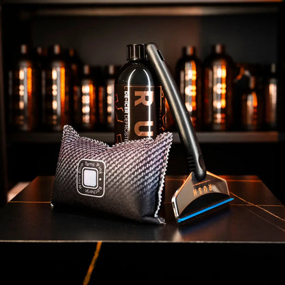 A bottle labeled RUSH from the Winter Essentials Package, a black and silver razor, and a black textured cleaning pad are shown on a dark surface with blurred De-Icer products in the background.