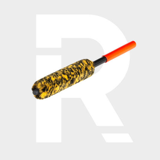 The Wheel Woolie Small is a cylindrical wheel brush with a black and yellow fuzzy head and an orange handle, shown on a light gray background with a large white "R"—ideal for cleaning spoke wheels.