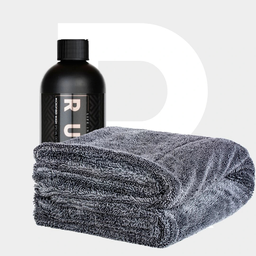 Een zwarte fles Microfiber Wash staat naast een netjes gevouwen grijze handdoek op een lichte achtergrond.