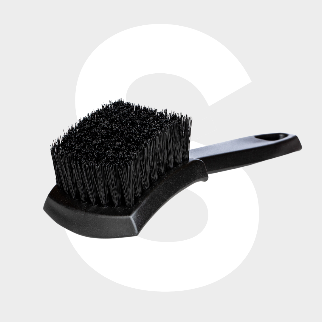 The Tire Brush, featuring a black design with stiff bristles and a curved handle for optimal banden reinigen, is shown on a light gray background with a large white letter S behind it.