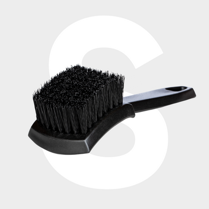 The Tire Brush, featuring a black design with stiff bristles and a curved handle for optimal banden reinigen, is shown on a light gray background with a large white letter S behind it.