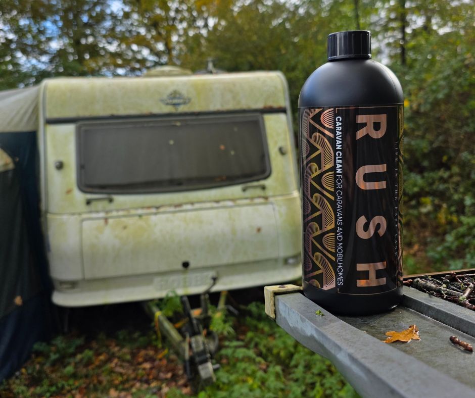 Een zwarte fles met het label RUSH staat buiten, perfect voor de caravanreiger, met een verweerde caravan en bomen zachtjes wazig op de achtergrond.