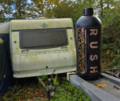 Een zwarte fles met het label RUSH staat buiten, perfect voor de caravanreiger, met een verweerde caravan en bomen zachtjes wazig op de achtergrond.