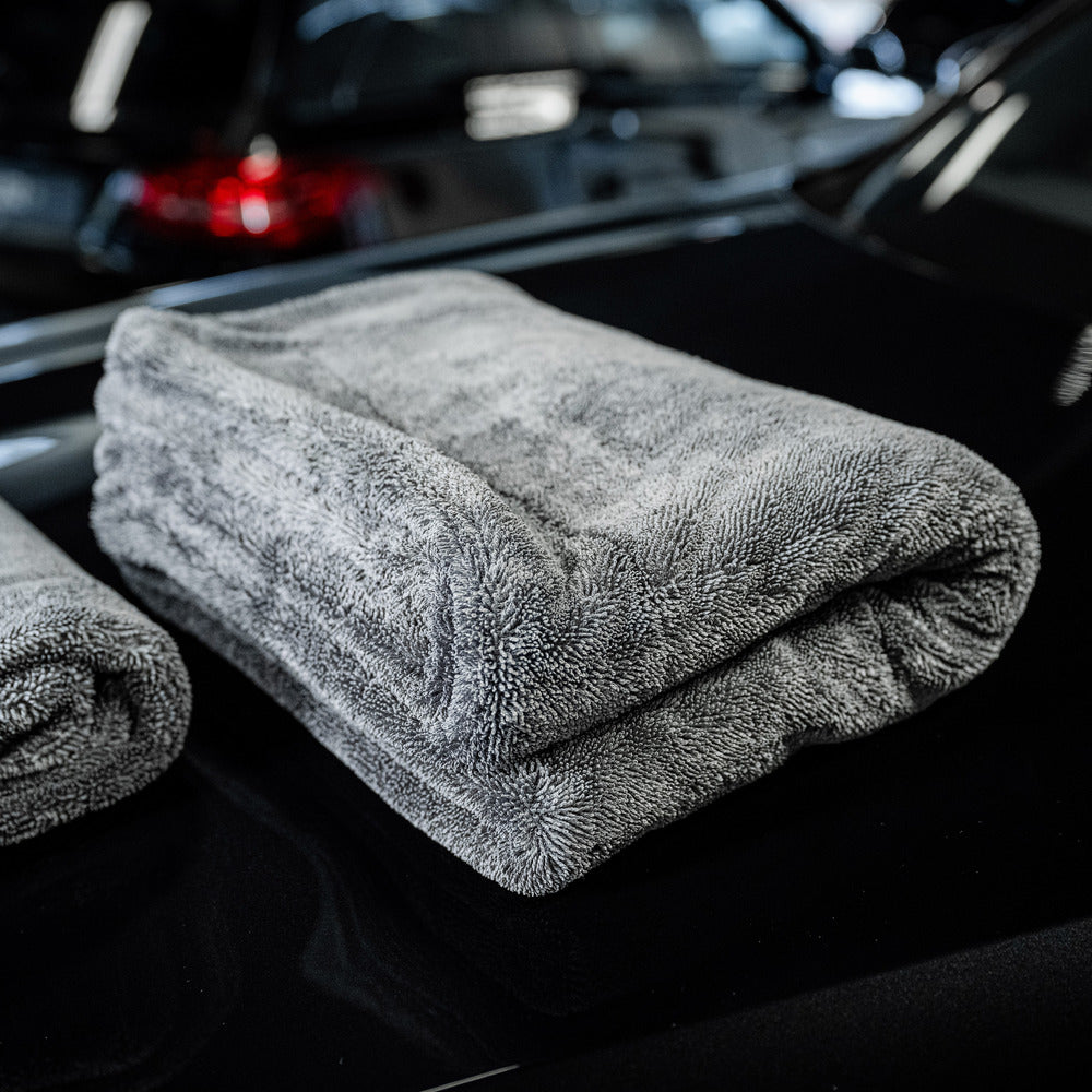 A large, folded Master Drying Towel 1600 GSM XL lies on a shiny black car surface, with another towel partially visible beside it and blurred cars in the background.
