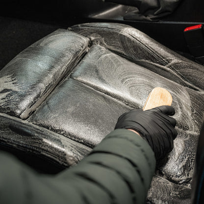 A person in black gloves uses a brush and Leather Matt Clean to scrub a leather car seat with foam, leaving the leather upholstery spotless and with a matte finish.