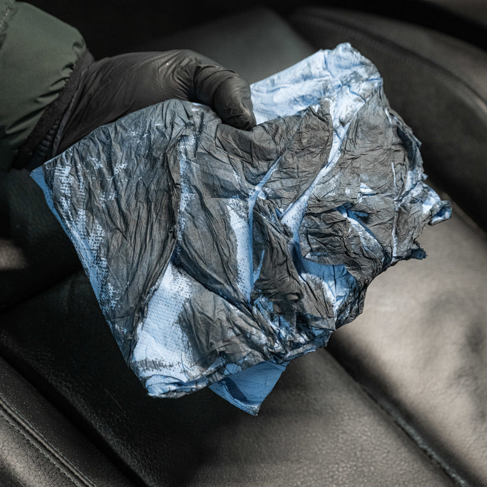 A hand in a black glove holds a wrinkled blue cloth over a black leather car seat, ready to use Leather Matt Clean for cleaning leather upholstery and achieving a matte finish.