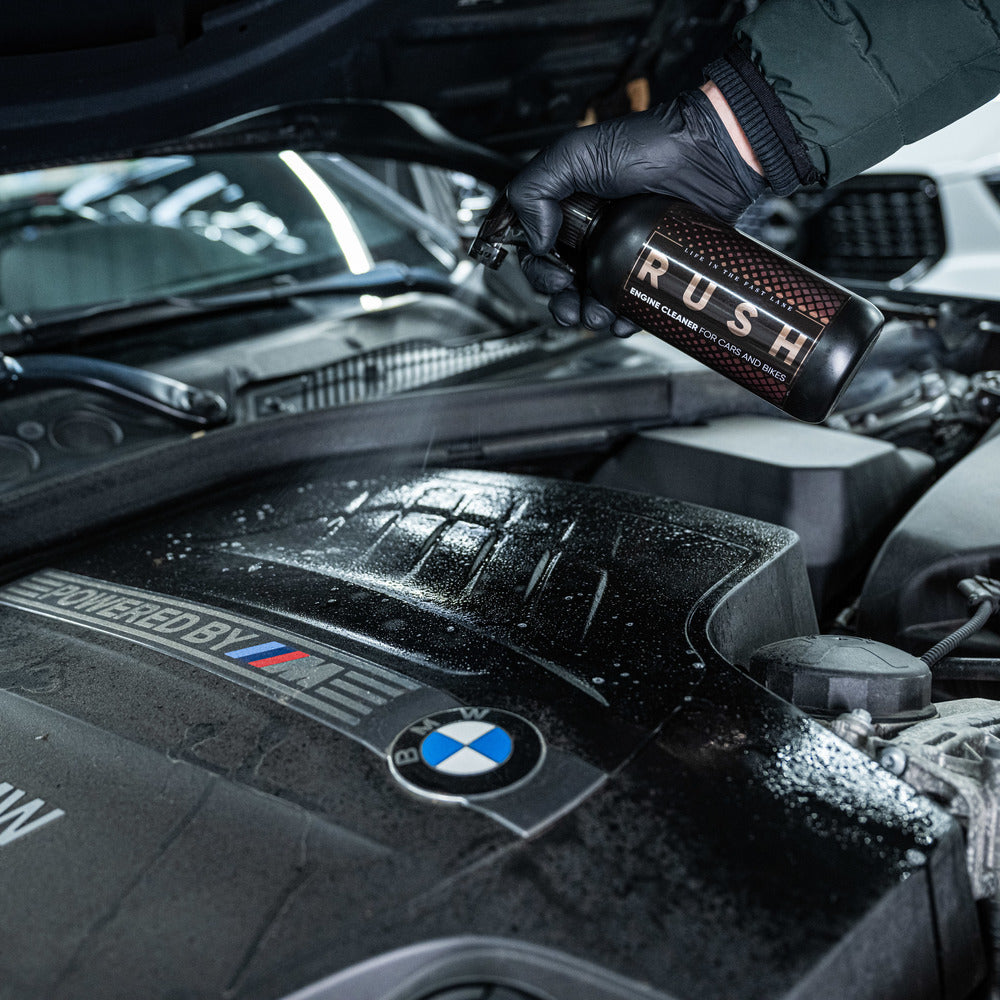 Een gehandschoende hand spuit RUSH Engine Cleaner, een krachtige alkalische reiniger, op de motorruimte van een BMW.
