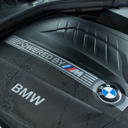 Close-up van een BMW motordeksel, ideaal voor motorruimte reiniging met RUSH Engine Cleaner; Powered by M en BMW logo zichtbaar.