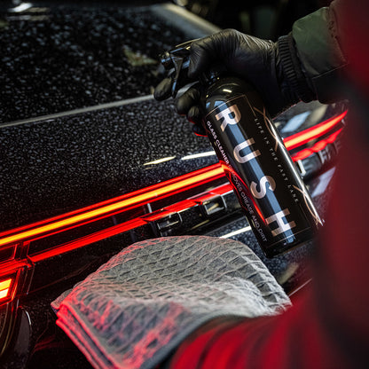 Een gehandschoende hand spuit RUSH glasreiniger op een zwarte auto in de buurt van zijn rode achterlicht, met een doek ernaast voor streepvrij glas reinigen.