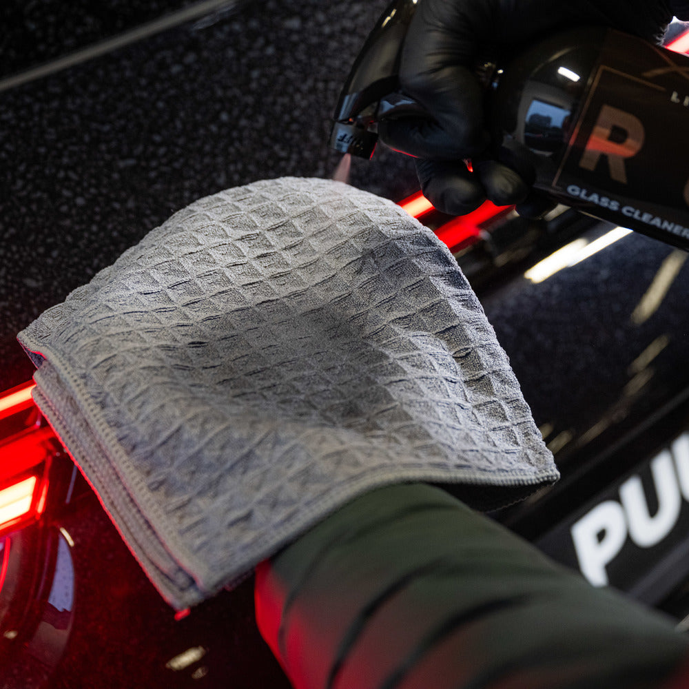 Een hand spuit glasreiniger op een gevouwen grijze structuurdoek bij het achterlicht van een zwarte auto voor streepvrij glas reinigen.