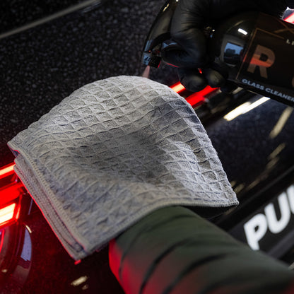 Een hand spuit glasreiniger op een gevouwen grijze structuurdoek bij het achterlicht van een zwarte auto voor streepvrij glas reinigen.