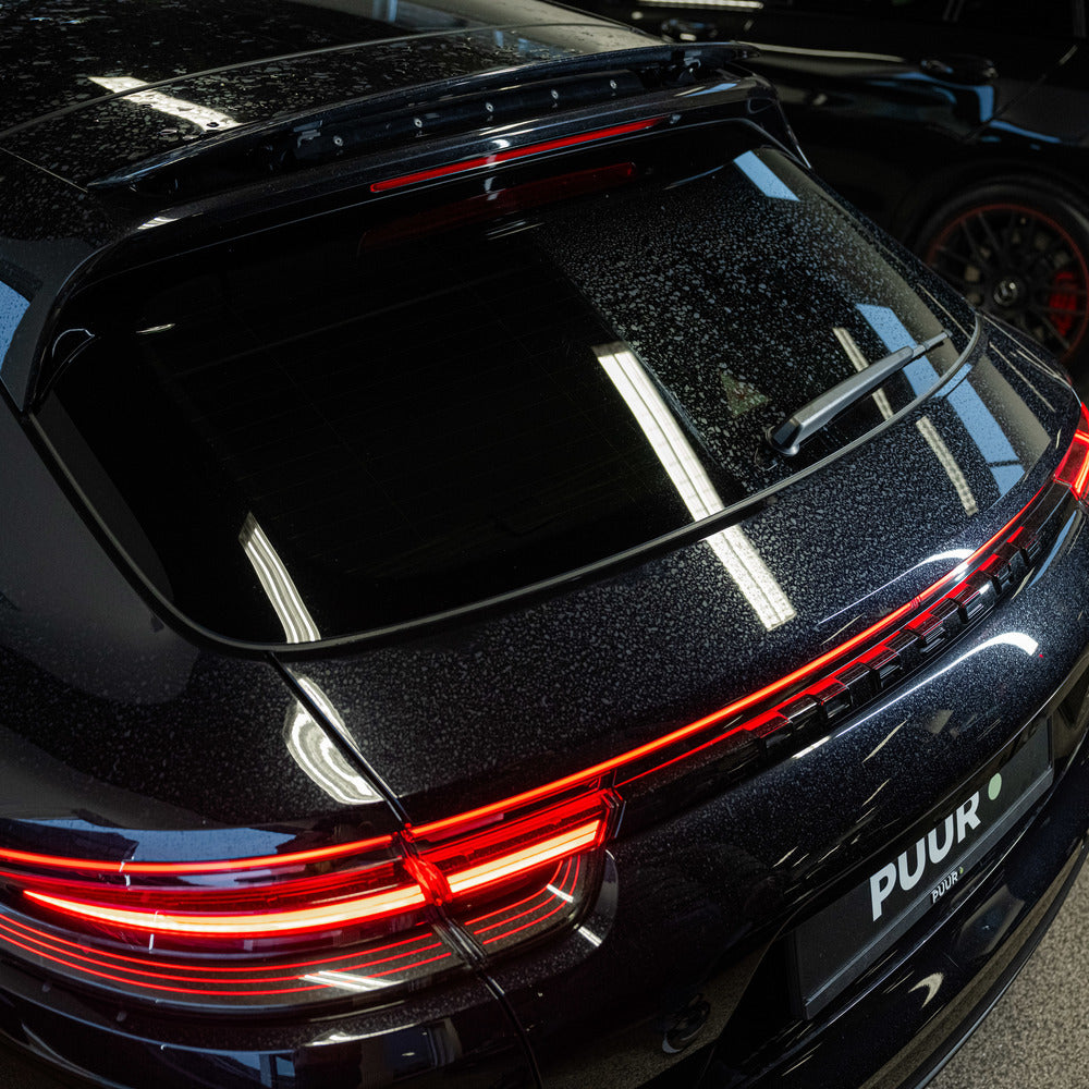 Close-up van de achterkant van een zwarte Porsche met streepvrij glas reinigen, verlichte achterlichten en een PUUR-kentekenplaat binnen onder felle lampen.