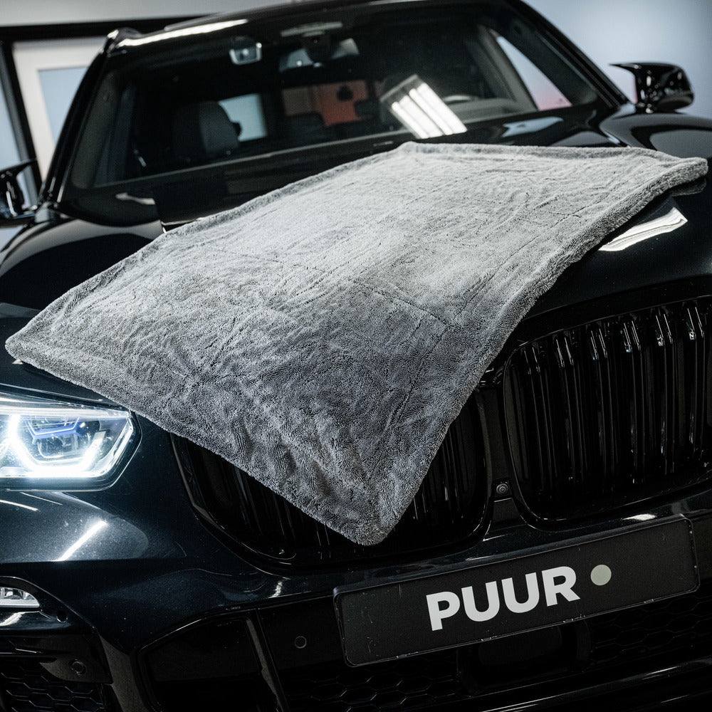 A Master Drying Towel 1600 GSM XL is draped over the hood of a shiny black car with lit headlights; the car's front grille and PUUR license plate are visible.