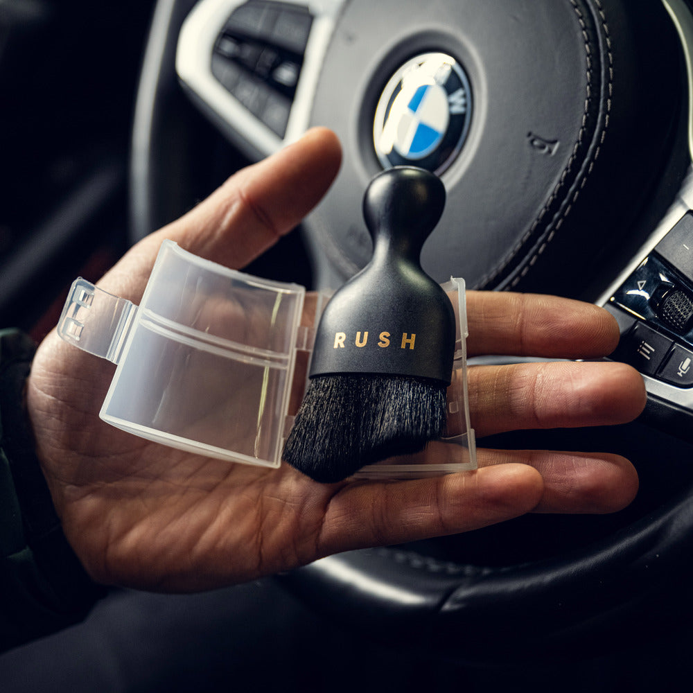 Een hand houdt een ultrazachte interieurborstel met het label RUSH met nanovezel borstelharen vast voor een BMW stuurwiel, gedeeltelijk in de koffer.