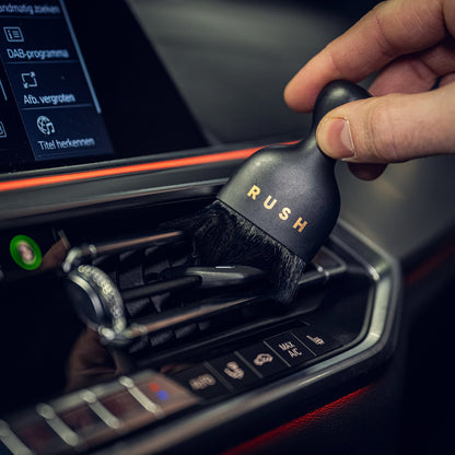 Een hand gebruikt een ultrazachte interieurborstel met het label RUSH om de dashboardbediening van de auto te reinigen met fijne nanovezel borstelharen.