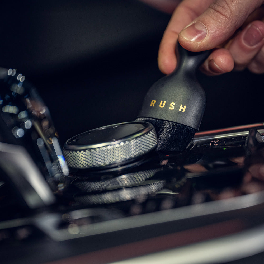 Een hand gebruikt een ultrazachte interieurborstel van het merk RUSH om een zilveren wijzerplaat op het glanzende dashboard van een auto schoon te maken.