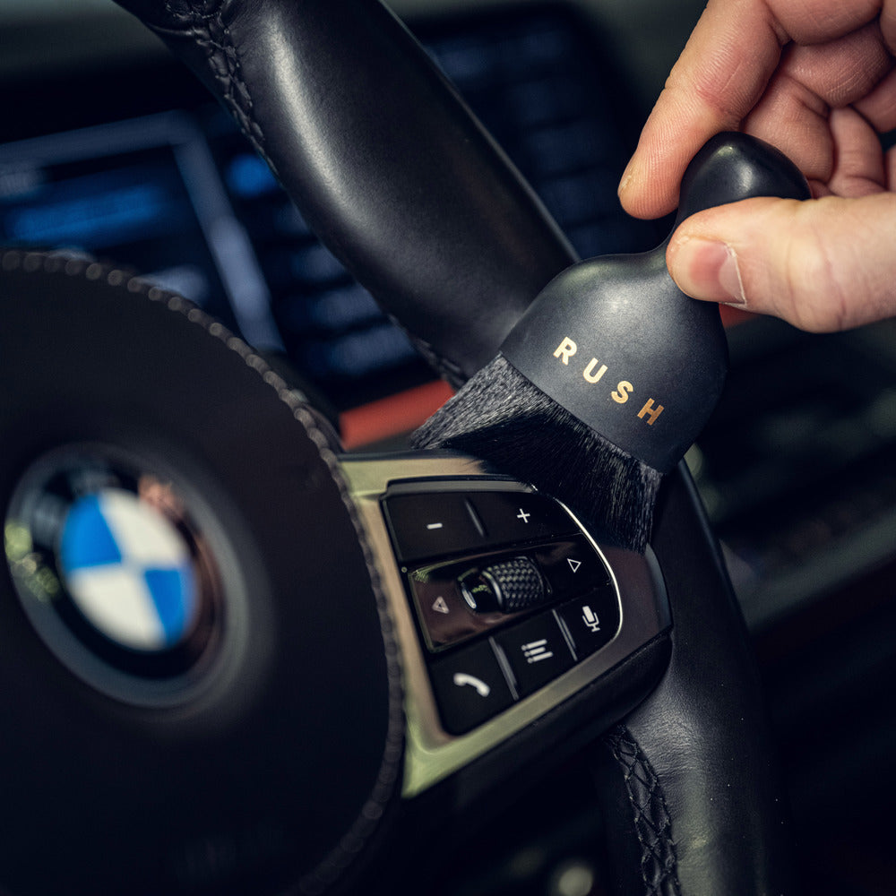 Een hand gebruikt een ultrazachte interieurborstel met nanovezel borstelharen om de bedieningselementen van het BMW stuurwiel in een auto schoon te maken.