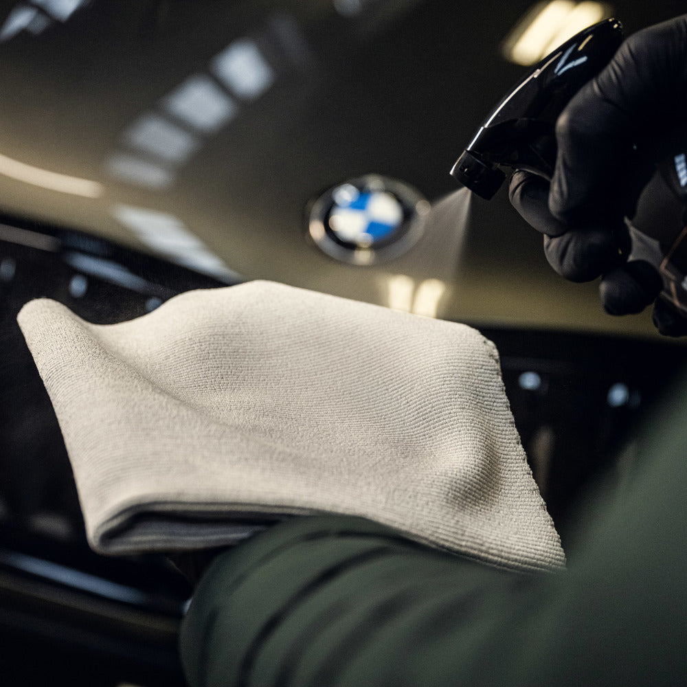 Een gehandschoende hand spuit reinigingsmiddel op een microvezeldoek voor de motorkap van een BMW, een demonstratie van vakkundig autodetailing.
