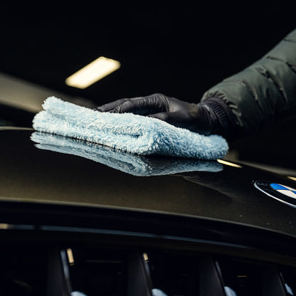 Een gehandschoende hand veegt de motorkap van een BMW schoon met RUSH Spray Wax, waardoor een beschermende laag ontstaat en de auto laat glanzen.