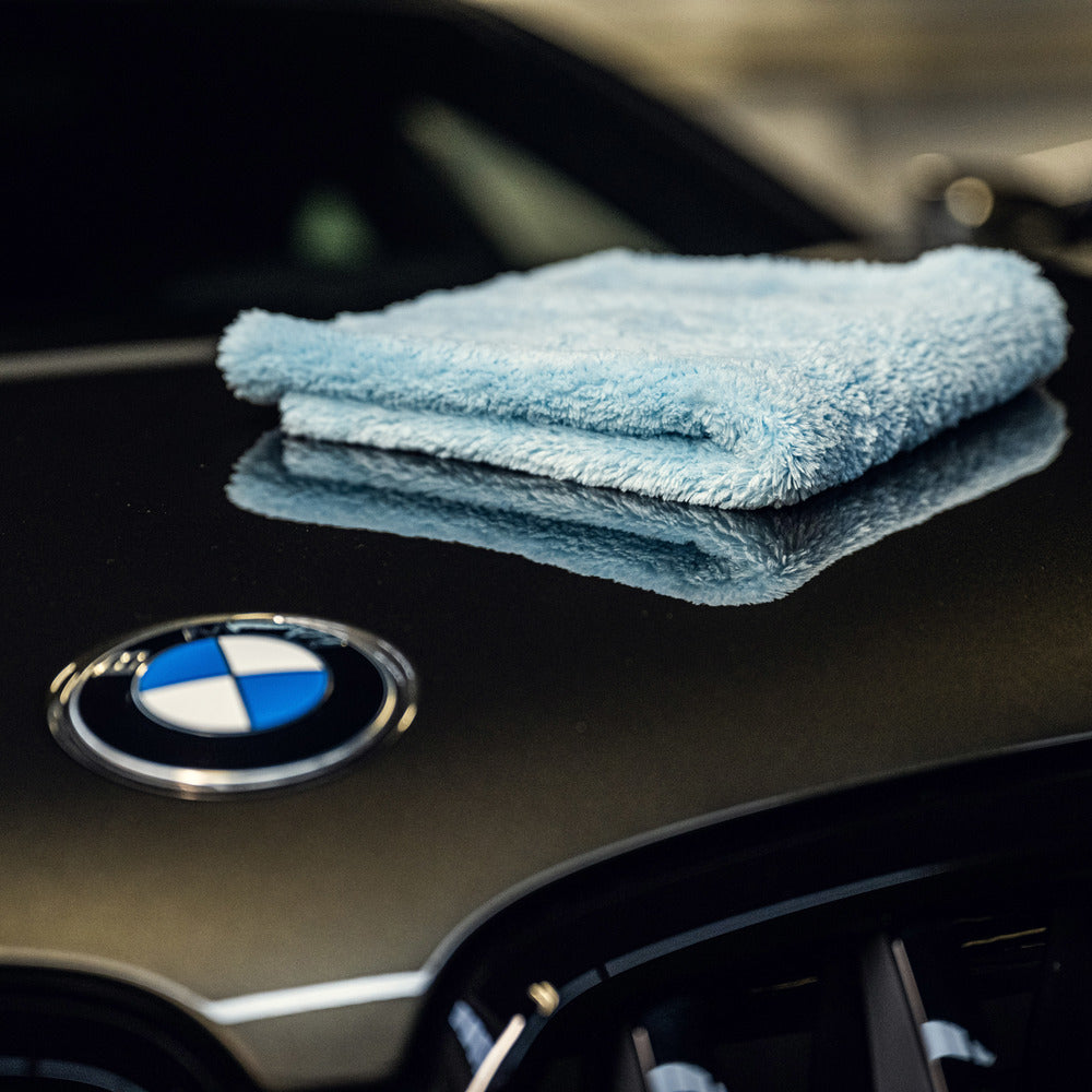 Een gevouwen blauw ultrazachte microvezeldoek, 500 GSM fleece, rust op de zwarte motorkap van een auto met een zichtbaar BMW embleem.