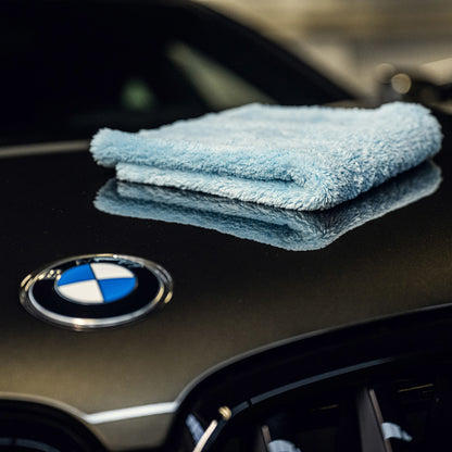 Een gevouwen blauw ultrazachte microvezeldoek, 500 GSM fleece, rust op de zwarte motorkap van een auto met een zichtbaar BMW embleem.