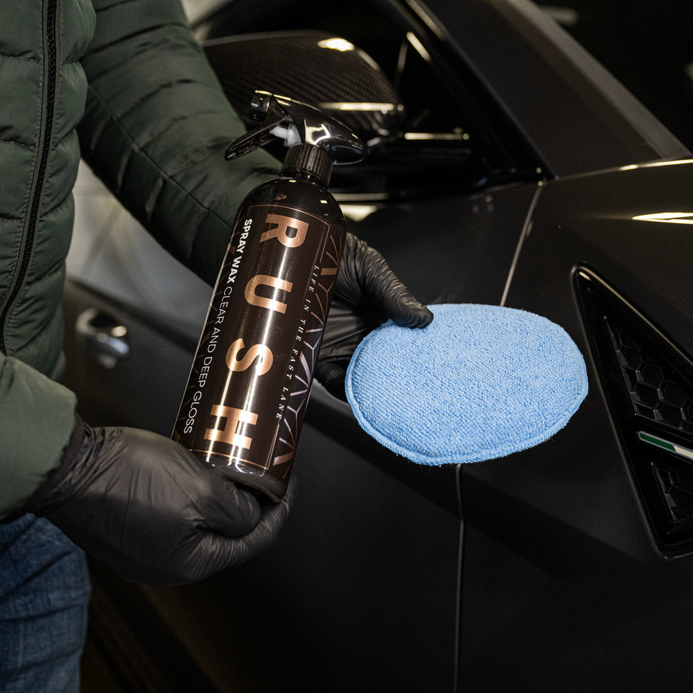 Een persoon met zwarte handschoenen houdt een fles RUSH en een autowaspad vast naast een zwarte autodeur.
