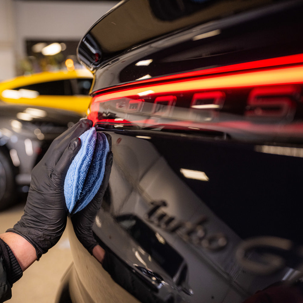 Een persoon met een zwarte handschoen gebruikt een applicatorpad en een blauwe microvezeldoek om het achterlicht van een zwarte auto schoon te maken.