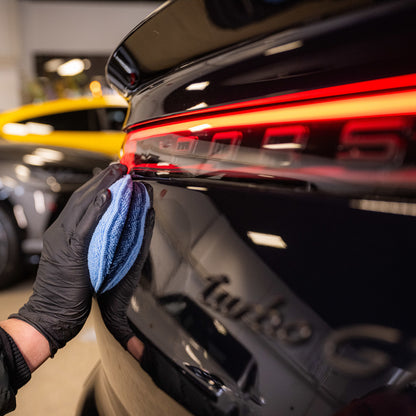 Een persoon met een zwarte handschoen gebruikt een applicatorpad en een blauwe microvezeldoek om het achterlicht van een zwarte auto schoon te maken.
