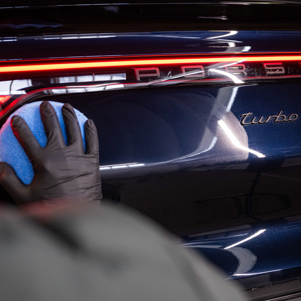 Een gehandschoende hand gebruikt een blauwe spons om de achterkant van een donkere Porsche Turbo te polijsten, waarbij de rode achterlichten zichtbaar zijn.