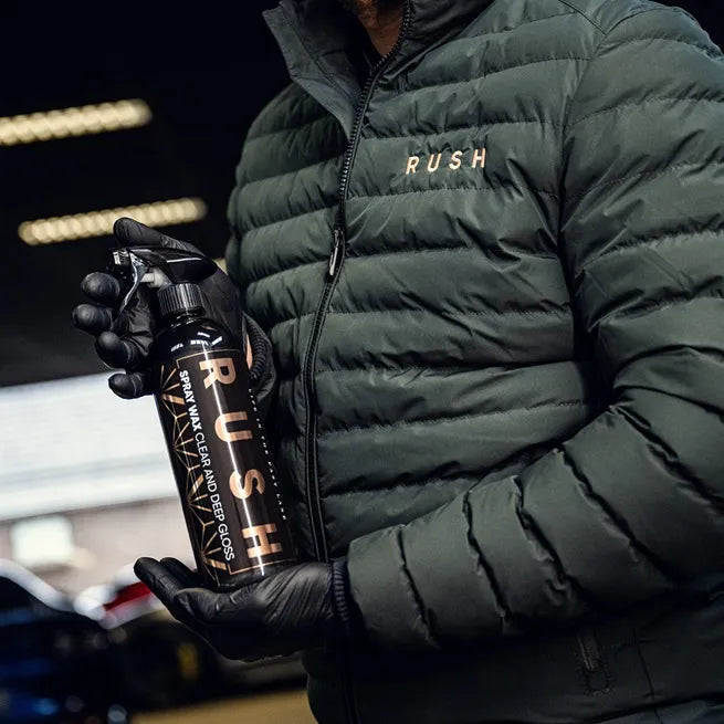 A person in a dark green puffer jacket and black gloves holds a black RUSH spray bottle, likely from the Detailing Package PLUS | 23-delig set, in a garage or workshop setting.