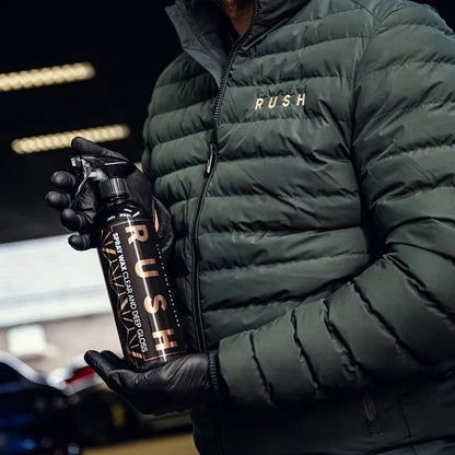 A person in a dark green puffer jacket and black gloves holds a black RUSH spray bottle, likely from the Detailing Package PLUS | 23-delig set, in a garage or workshop setting.