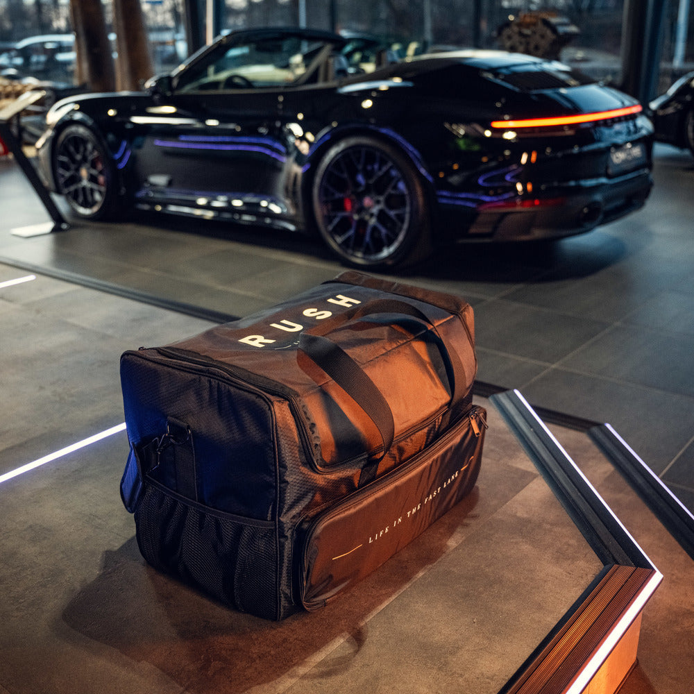 Een zwarte RUSH Detailing Bag, een ruime opbergtas voor schoonmaakproducten opbergen, staat binnen met een zwarte sportauto op de achtergrond.